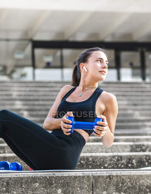 Young sportswoman looking and and listening to music while doing abs exercise with dumbbell on stairs during fitness workout on city street — Stock Photo