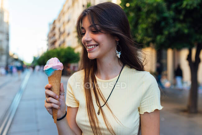 Растениеводная молодая женщина в кулоне и серьги с вкусным мороженым в вафельном конусе глядя на улицу — стоковое фото