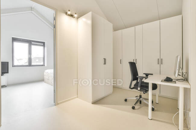 Comoda sedia e tavolo con computer posizionato vicino all'armadio in stile moderno minimalista mansarda con design interno bianco — Foto stock
