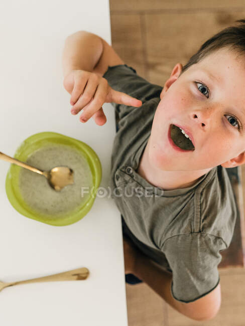 Вид сверху на очаровательного мальчика, сидящего за столом с полным ртом во время обеда дома — стоковое фото