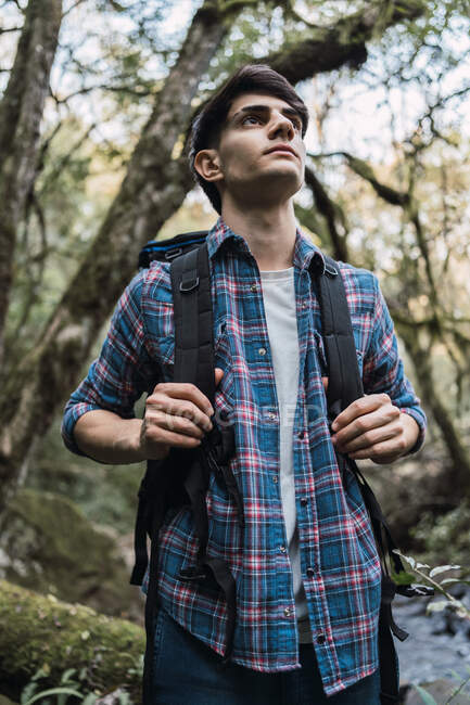 Vista lateral do homem viajante com mochila em pé na estrada arenosa na floresta durante o trekking e olhando para longe — Fotografia de Stock