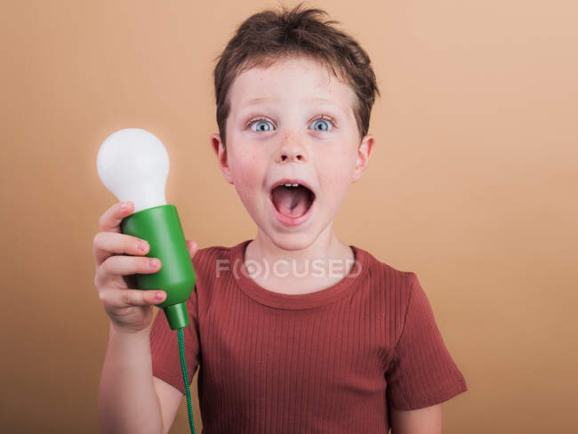 Дивовижна дитина в сорочці з пластиковою лампочкою, що представляє ідею, дивлячись на камеру на бежевому фоні — стокове фото