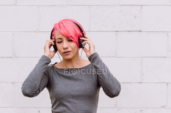 Mujer joven con pelo rosa brillante escuchando música con auriculares mientras está de pie cerca de la pared blanca con los ojos cerrados - foto de stock