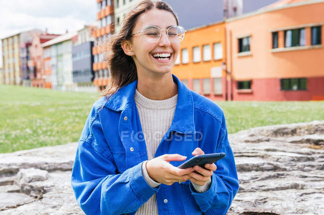 Positive young female in stylish clothes standing in city street and messaging on mobile phone while laughing with closed eyes — Stock Photo
