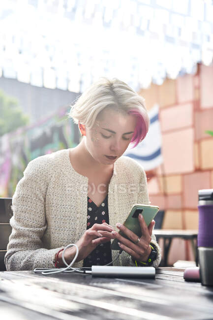 Альтернативная женщина с короткими волосами, просматривающая социальные сети на смартфоне, сидя за столом в уличном кафе в солнечный день — стоковое фото