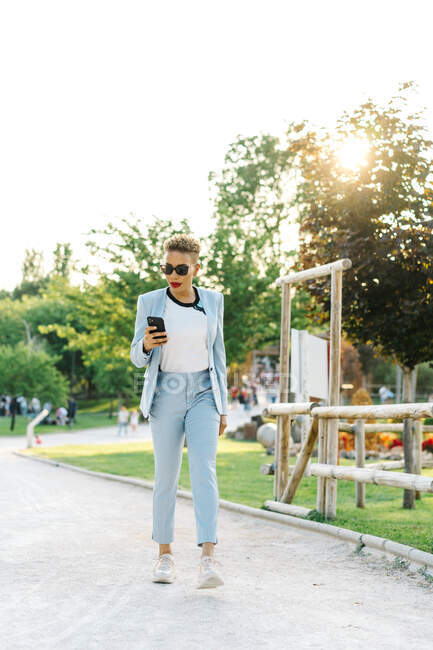 Stylish African American female in sunglasses text messaging on cellphone while strolling on walkway in urban park — Stock Photo