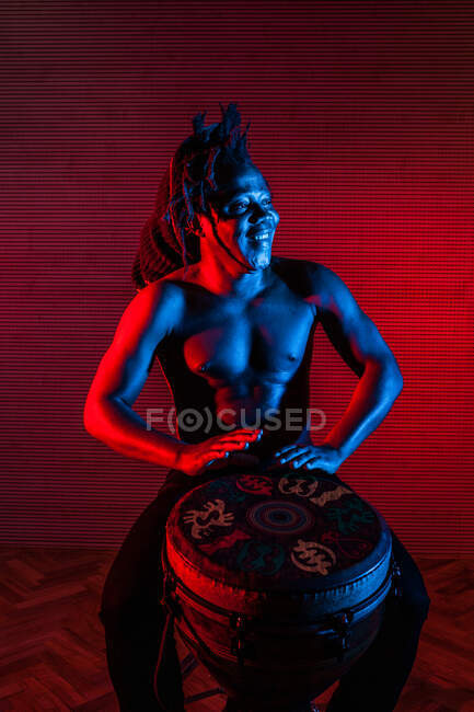 Musicien masculin noir rêveur avec torse nu jouant du tambour africain en studio avec des néons rouges et bleus — Photo de stock