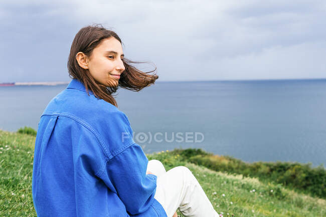 Vista laterale della deliziosa femmina seduta sulla collina verde e godendo della vista sul mare nella giornata nuvolosa — Foto stock