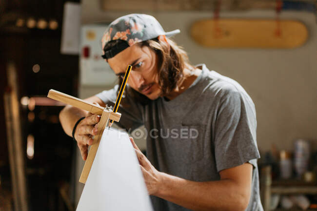 Hipster masculino concentrado usando herramienta de escriba con lápiz mientras marca la tabla de surf antes de dar forma en el taller - foto de stock