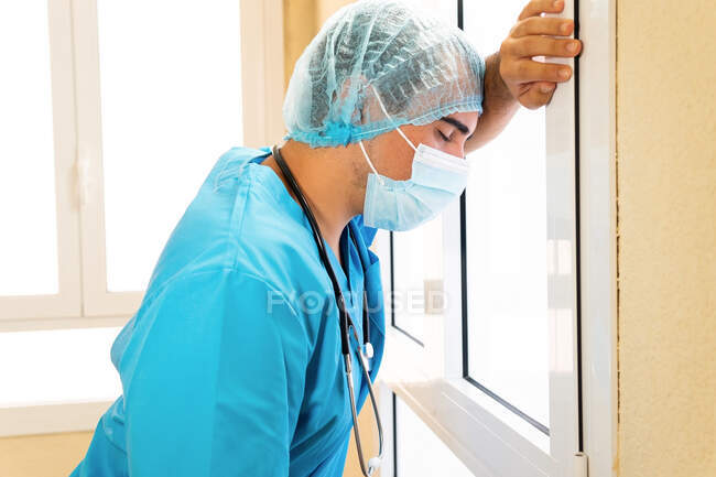 Vista lateral del médico agotado en uniforme y máscara protectora de pie cerca de la pared después de un duro día de trabajo en la clínica durante COVID - foto de stock