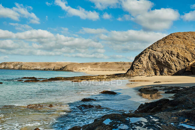 Широкий кут зору на мирну лагуну з чистою морською водою біля піщаного пляжу і скелями проти хмарного сходу неба в Фуертевентурі (Іспанія). — стокове фото