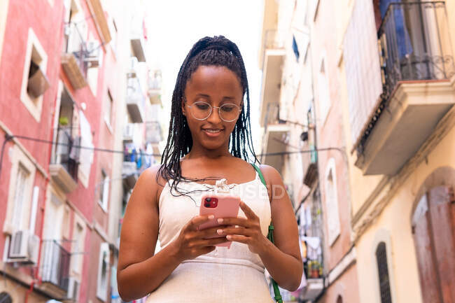 Низький кут посмішки афро-американської жінки з брамами повідомлень в соціальних мережах через мобільний телефон, стоячи на вулиці з старими будівлями в Барселоні. — стокове фото