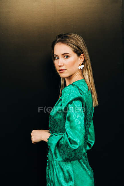 Vista laterale di donna serena in abito verde alla moda in piedi su sfondo marrone e guardando la fotocamera — Foto stock