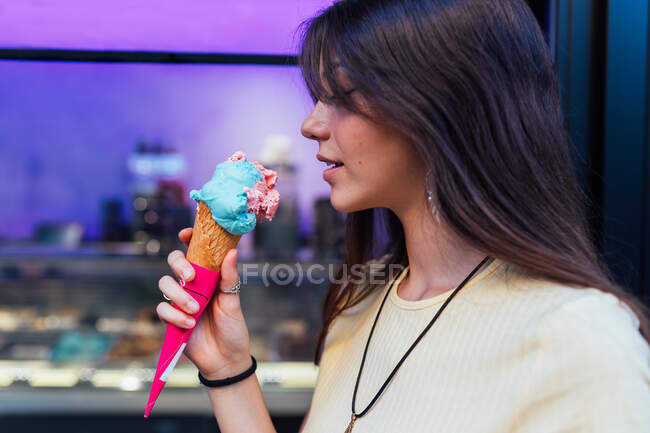Cosecha alegre joven hembra en colgante y pendientes con delicioso helado en cono de gofre mirando hacia la calle - foto de stock