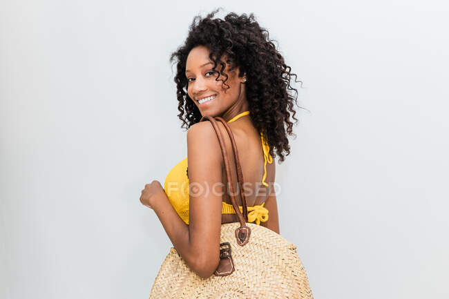 Back view of cheerful ethnic woman with wicker bag looking at camera over shoulder on light background — Stock Photo