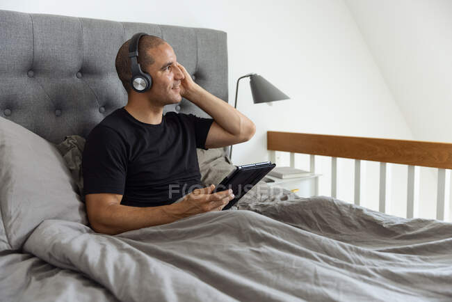 Sereno macho em fones de ouvido sentado na cama sob cobertor e navegando nas mídias sociais em tablet enquanto ouve música de manhã — Fotografia de Stock