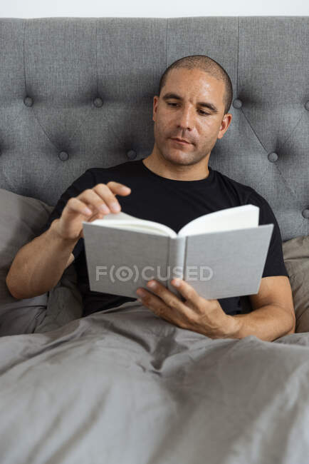 Pensivo macho sentado em cama macia de manhã e lendo história interessante no livro após o despertar — Fotografia de Stock