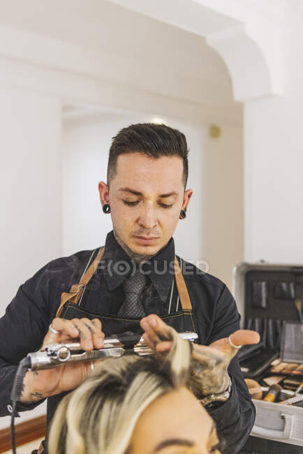 Мужчина-парикмахер, использующий утюг для завивки блондинистых замков женщины-клиента во время работы в салоне красоты — стоковое фото