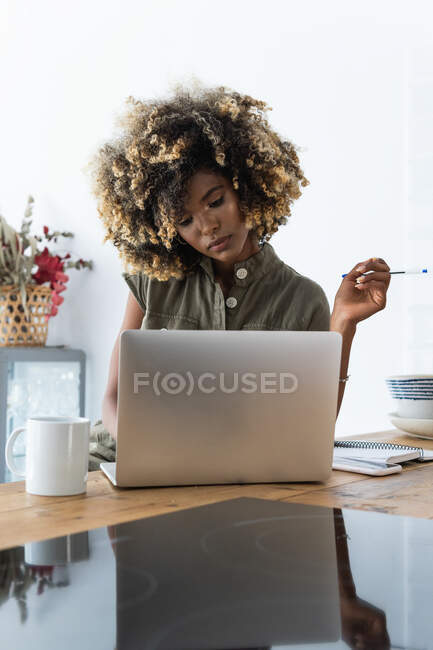Freelancer afroamericana concentrada escribiendo en bloc de notas mientras está sentada en la mesa con el portátil y trabajando remotamente en casa - foto de stock