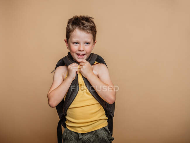 Positive cool preteen schoolboy with rucksack looking away on brown background in studio — Stock Photo