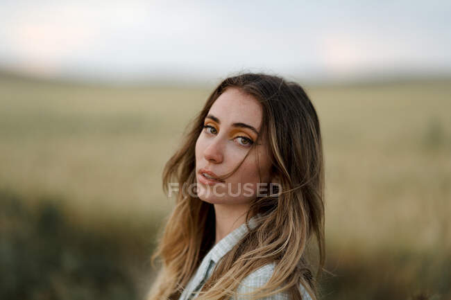Side view of young mindful female looking at camera on road near meadow under cloudy sky in evening in countryside — Stock Photo