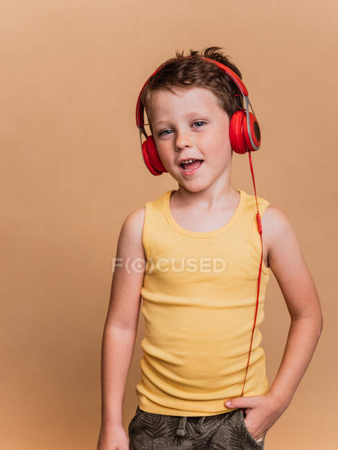 Menino pré-adolescente satisfeito em fones de ouvido vermelhos ouvindo música e assinando música em fundo marrom em estúdio — Fotografia de Stock