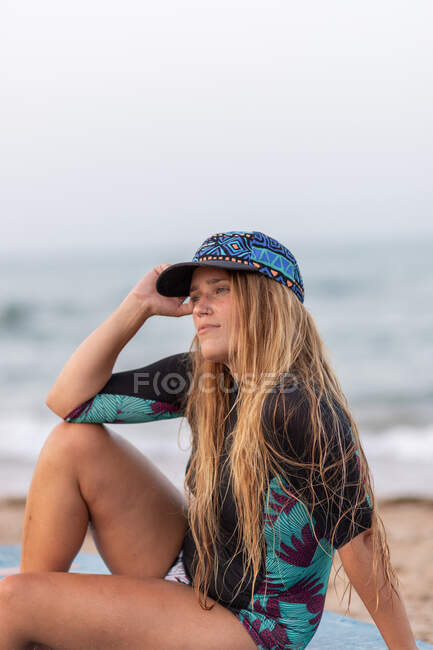 Side view of thoughtful female surfer in swimwear and hat sitting on paddleboard on sandy shore against sea and looking away — Stock Photo