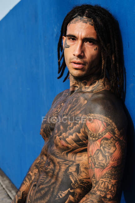 Hombre étnico de moda sin camisa con trenzas y tatuajes de pie cerca de la pared azul en la calle y mirando a la cámara - foto de stock