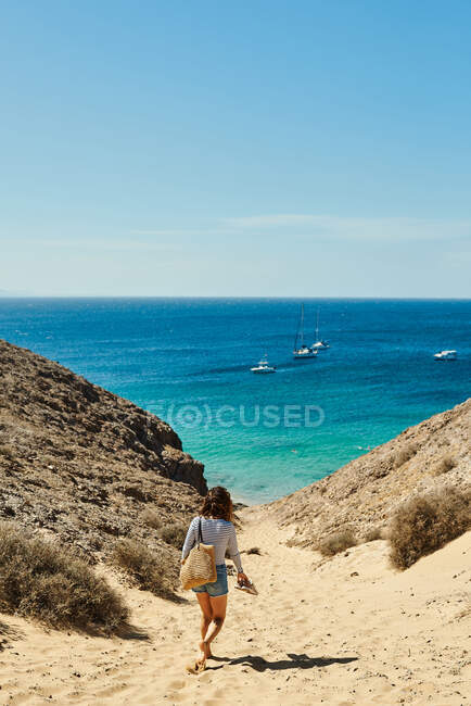 Vista trasera de la mujer en ropa casual mirando hacia otro lado y admirando el mar turquesa mientras camina por la costa arenosa durante las vacaciones de verano en Fuerteventura, España - foto de stock