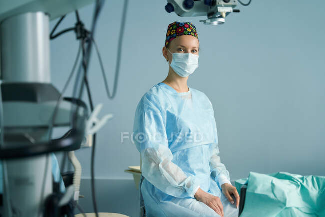 Doctora adulta en uniforme quirúrgico y máscara estéril mirando a la cámara mientras está sentada en la clínica - foto de stock