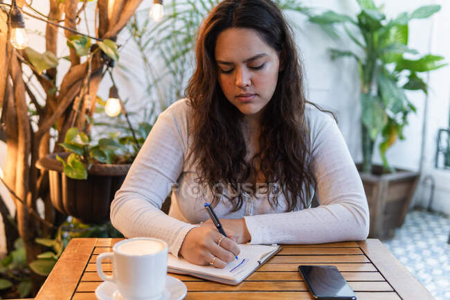Концентрована молода етнічна бізнес-леді в повсякденному одязі робить нотатки в планувальнику, сидячи за столом з чашкою кави і смартфона в затишній кафетерії з зеленими рослинами — стокове фото