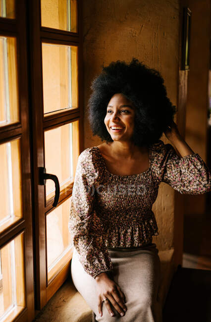 Encantada hembra afroamericana con el pelo rizado sentado en el alféizar de la ventana de madera en la casa y mirando hacia otro lado - foto de stock