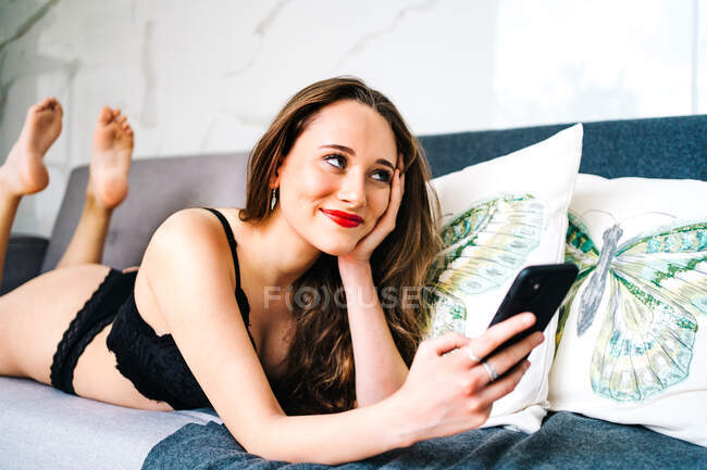 Mujer encantada en ropa interior negra tumbada en el sofá y mensajería en las redes sociales a través del teléfono móvil en el acogedor salón en casa - foto de stock