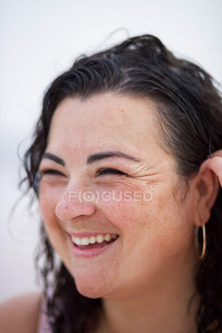 Усміхнена пишна жінка регулює вологе хвилясте волосся і дивиться в сторону розмитого фону — стокове фото