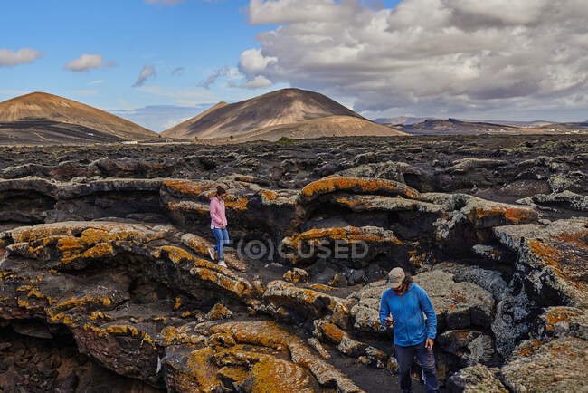 Desde arriba, hombre y mujer con mochilas caminando por la ladera de la montaña contra el cielo azul nublado en Fuerteventura, España - foto de stock