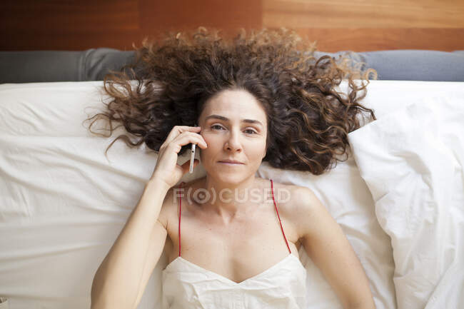 Вид сверху деловой женщины с кудрявыми волосами, лежащей в кровати и разговаривающей по телефону — стоковое фото
