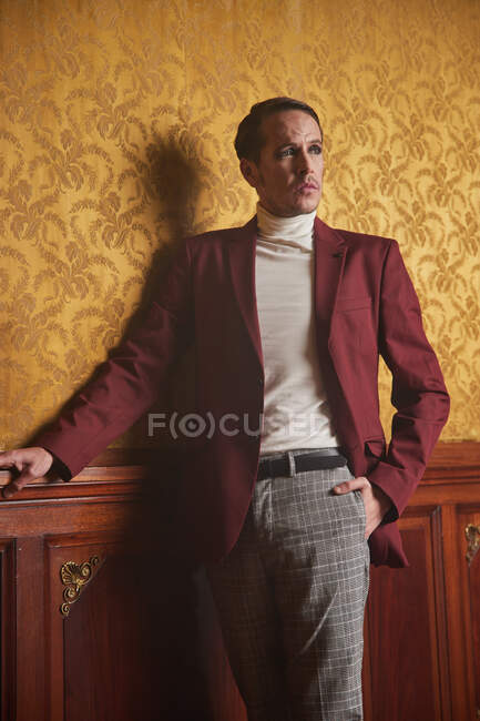 Впевнений дорослий чоловік-актор в елегантному стильному одязі тримає руку в кишені і задумливо дивиться, стоячи біля стіни в старовинному стилі кімнати — стокове фото
