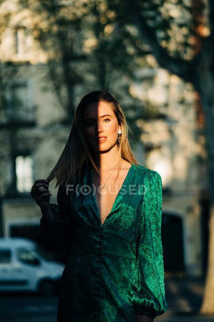 Femme insouciante en robe verte tendance debout avec une main dans les cheveux dans la rue et regardant la caméra tandis qu'un bâtiment l'ombrage — Photo de stock