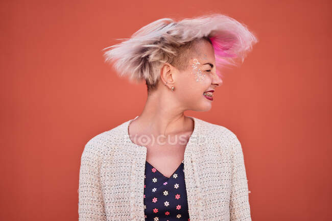 Despreocupado mujer alternativa lanzando cabellos cortos teñidos contra la pared naranja en el área urbana - foto de stock
