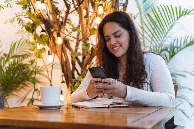 Mensajería femenina étnica joven en el teléfono móvil con bloc de notas mientras está sentado en la mesa con una taza de café y un portátil en la cafetería - foto de stock