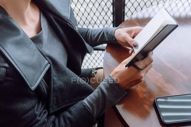 De arriba de la cosecha la hembra anónima en la ropa casual que sostiene ha abierto el cuaderno sobre la mesa de madera - foto de stock