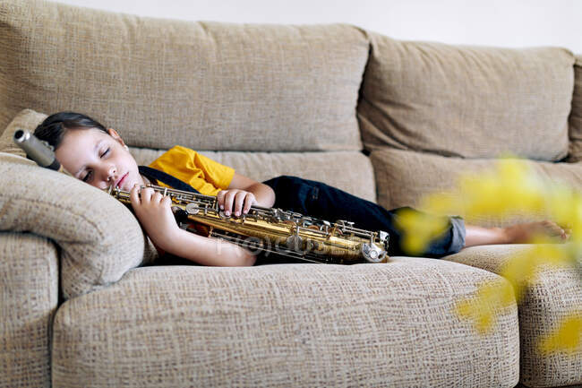 Очаровательный ребенок с саксофоном дремлет на диване в комнате — стоковое фото