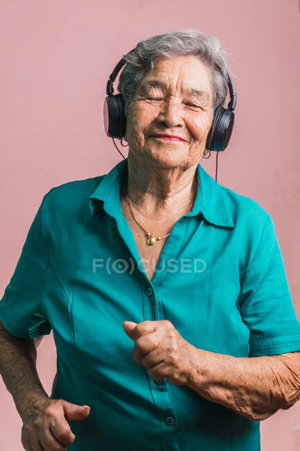 Joyful modern senior female listening to music in headphones and dancing with closed eyes on pink background in studio — Stock Photo