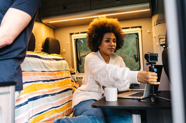 Young African American woman adjusting tripod with smartphone while sitting at table with laptop and preparing to record video for vlog during travel with boyfriend in camper van — Stock Photo
