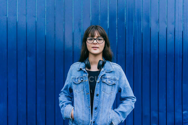 Charmante Frau in Jeansjacke und mit Kopfhörern am Hals steht auf blauem Hintergrund auf der Straße und schaut in die Kamera — Stockfoto