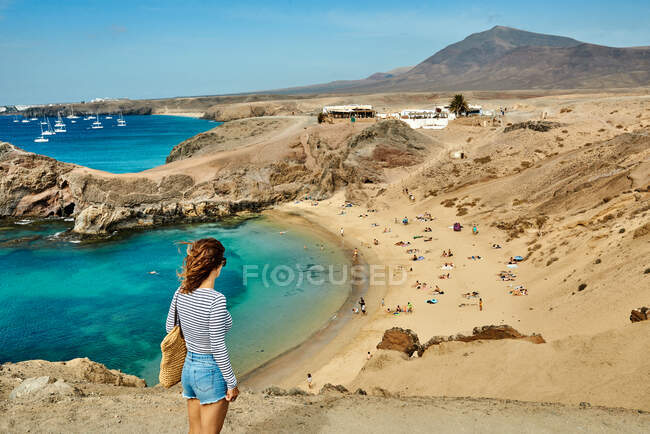 Visão traseira de turista feminina irreconhecível em roupas casuais que ficam perto de ruínas de pedra e atirando mar azul-turquesa no dia de verão em Fuerteventura, Espanha — Fotografia de Stock