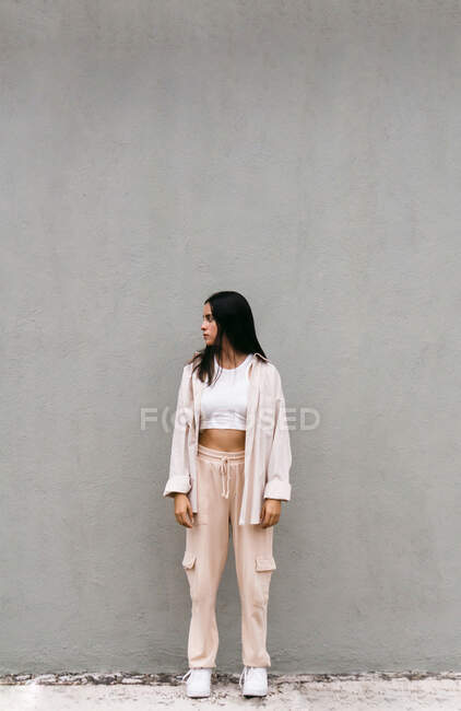Giovane donna in abito stile strada in piedi su sfondo di muro grigio in città e guardando altrove — Foto stock