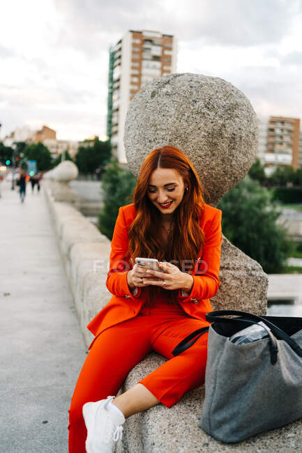 Happy stylish female with red hair and in vibrant orange suit sitting on stone border in city and messaging on cellphone — Stock Photo