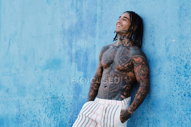 Hombre étnico de moda sin camisa con trenzas y tatuajes de pie cerca de la pared azul en la calle y los ojos cerrados - foto de stock