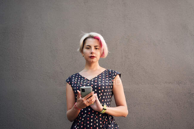 Entzückte ungezwungene Frau mit kurzen Haaren und im Kleid, die per Handy Nachrichten sendet, während sie auf der Straße vor einer grauen Wand steht und in die Kamera schaut — Stockfoto